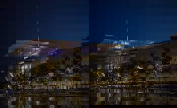 Photo taken at night showing a lake with trees around it and a large glass building behind it